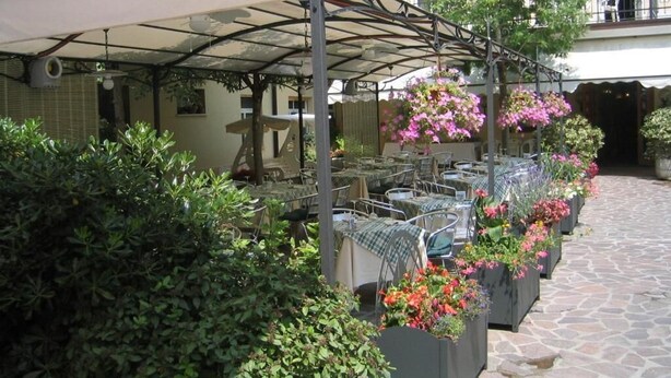 An outdoor café with tables and chairs under a covered area, surrounded by greenery and potted plants with flowers. - Hotel Europa (Viadana)
