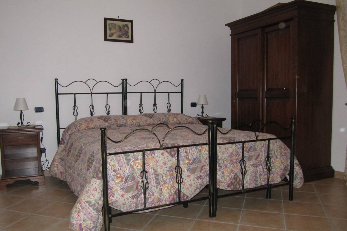 A bedroom with a bed, two bedside tables, a nightstand, a wooden wardrobe, and a wall-mounted picture.
