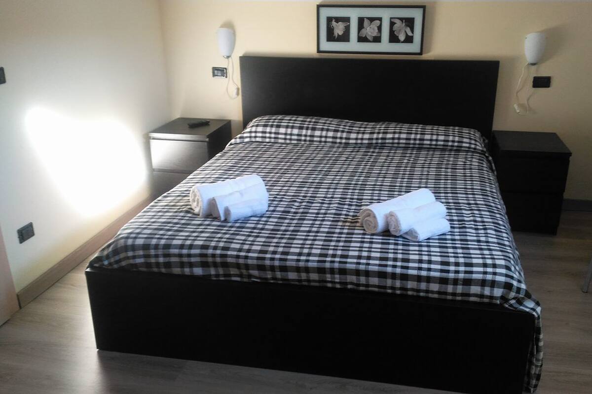 A neatly made bed with a checkered bedspread, two rolled towels, a bedside table with a lamp, and a chair.