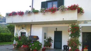 A two-story building with a white facade, green shutters, and a stone base. There are potted plants and flowers on the balconies, and a small table with chairs outside.