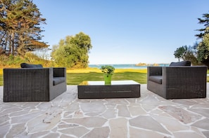 An outdoor seating area with wicker furniture and a stone floor, overlooking a grassy landscape and a body of water.