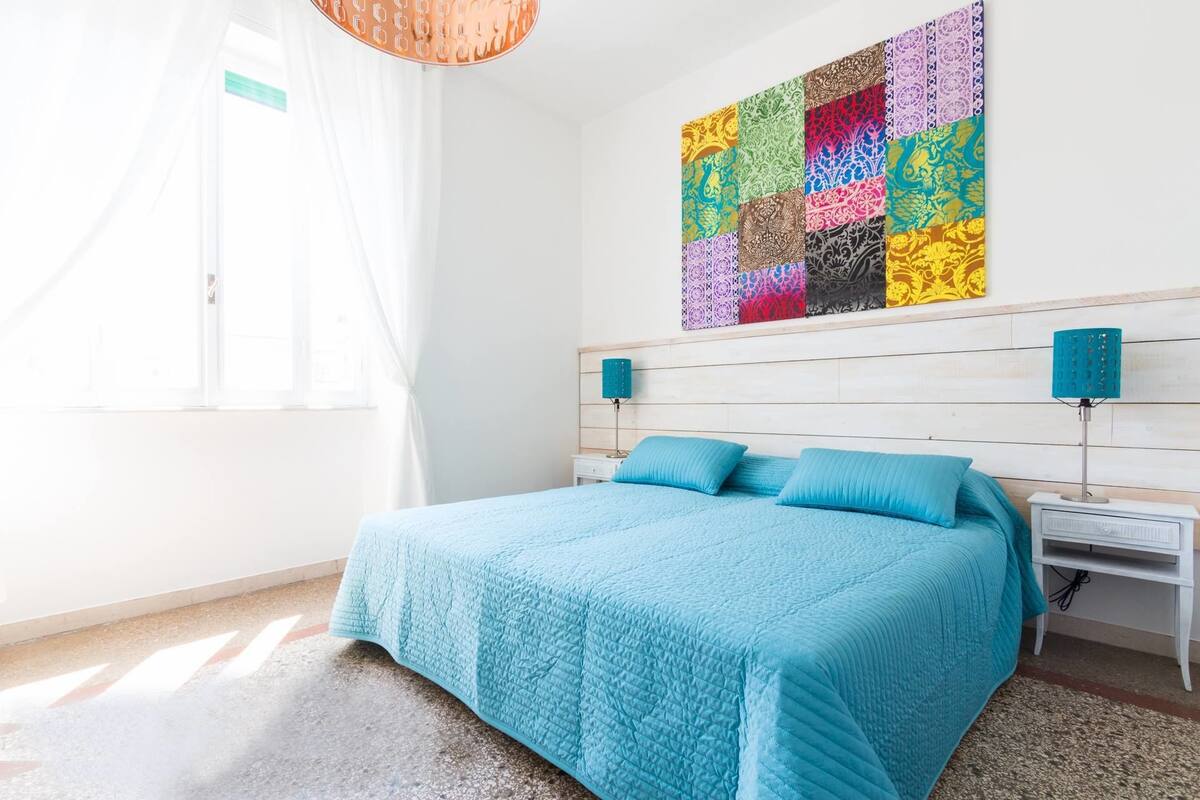Une chambre à coucher avec un lit, une œuvre d’art colorée accrochée au mur et une fenêtre avec des rideaux.