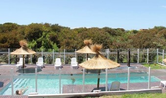 Una piscina con sillones de descanso y sombrillas de paja, rodeada por una terraza de madera y una valla de metal.