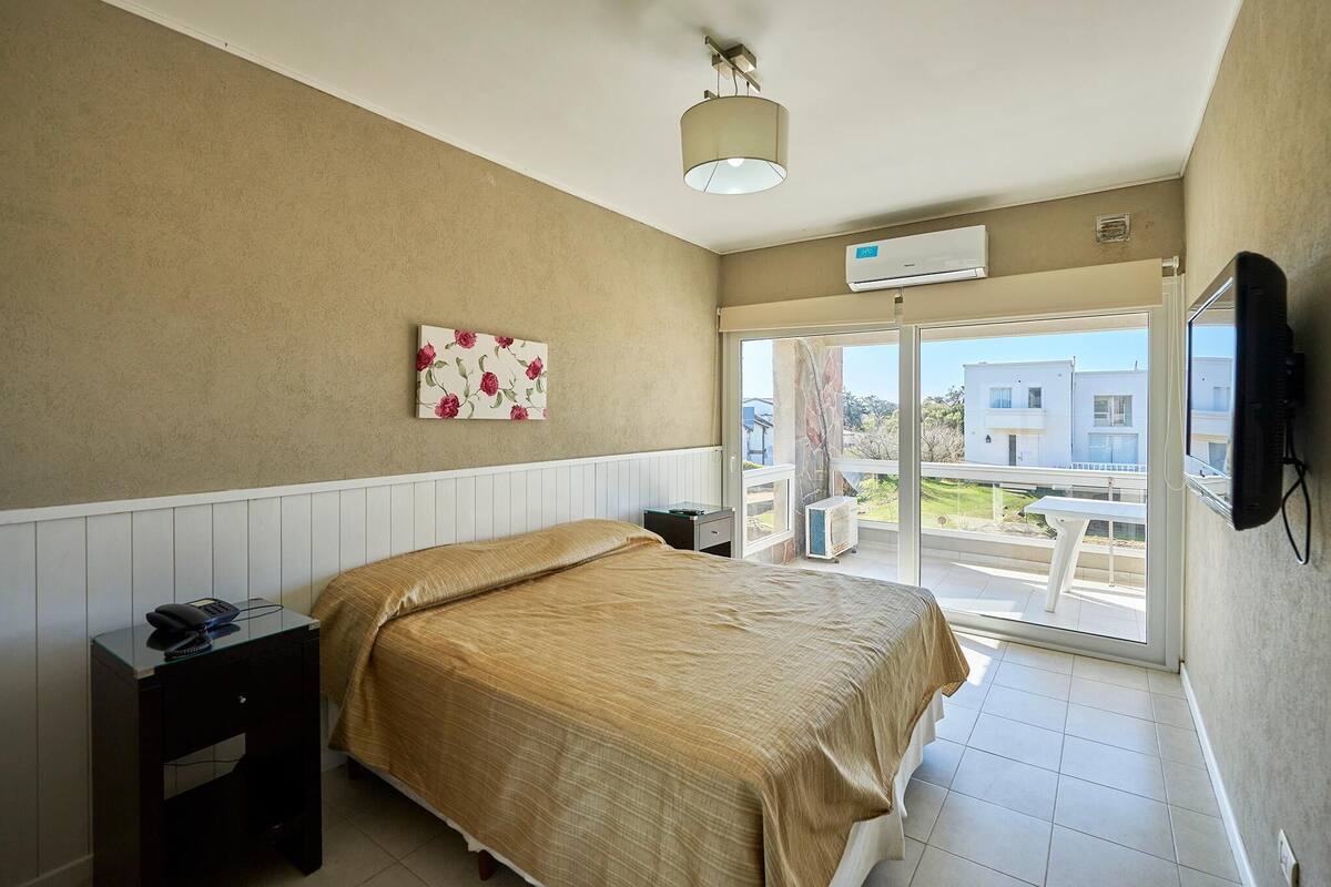 Una camera d'albergo con un letto, un comodino e un quadro appeso al muro. Si vede una finestra con vista sugli edifici e un balcone.