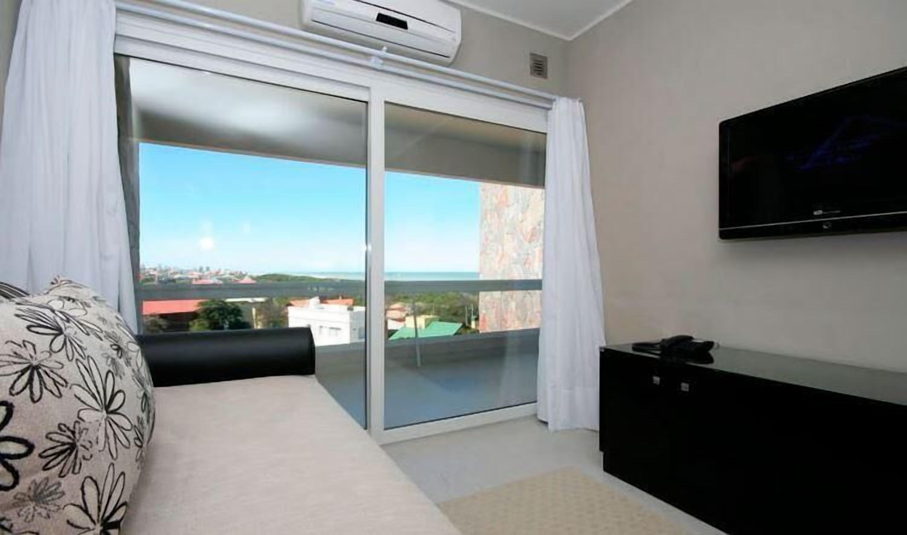 A bedroom with a bed, a TV, a sliding glass door to a balcony, and an air conditioning unit.