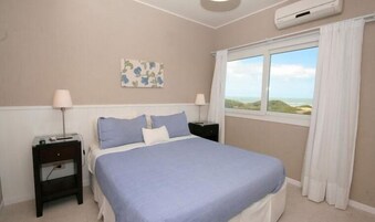 Un dormitorio con cama, dos mesitas de noche, aire acondicionado de pared, ventana con vista a la playa y una obra de arte floral en la pared.