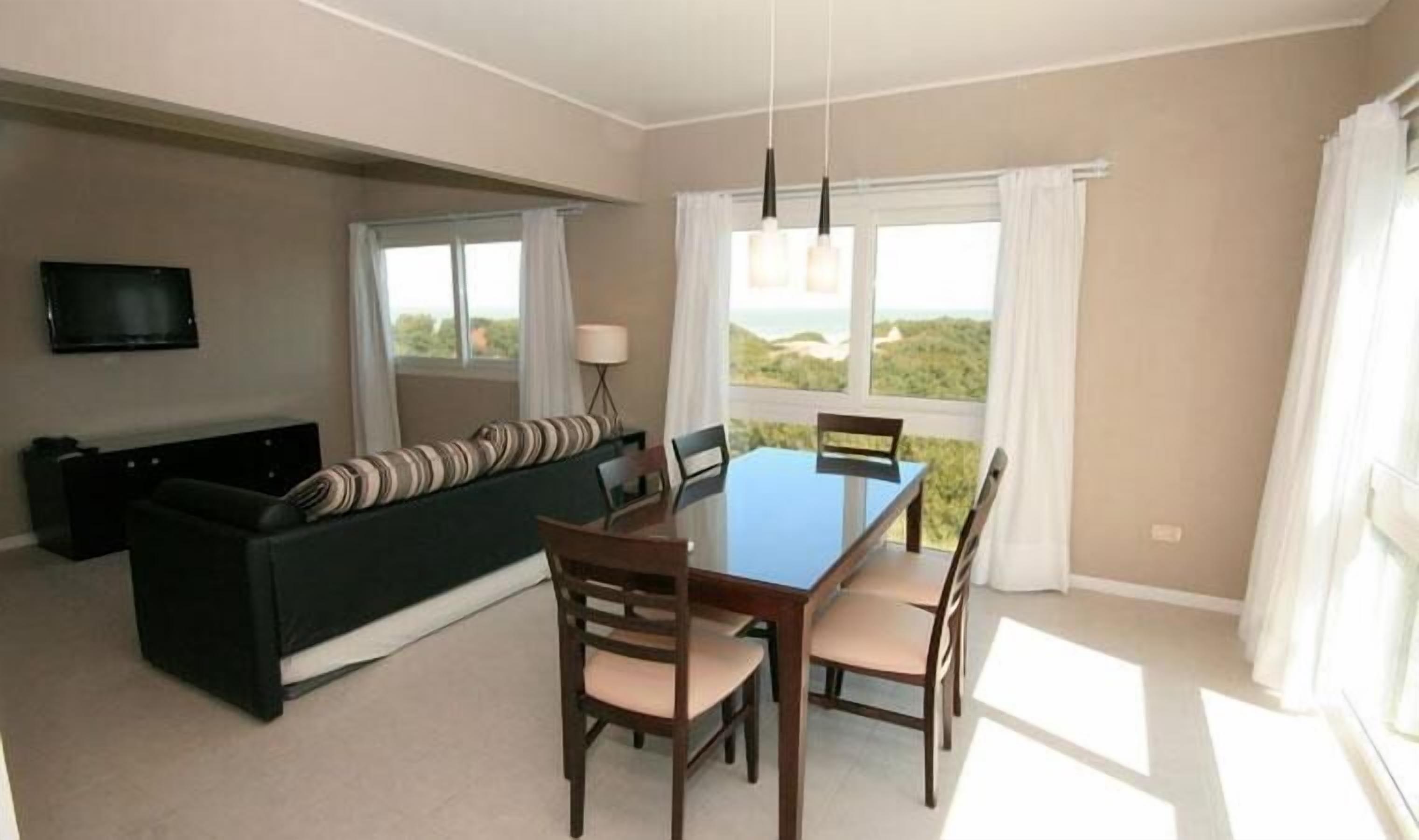 A modern living room with a dark green sofa, a wooden dining table with four chairs, a flat-screen TV mounted on the wall, and large windows with white curtains.