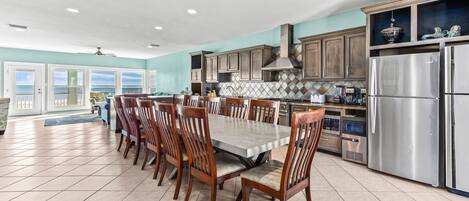 A spacious kitchen with a large dining table surrounded by chairs, a refrigerator, and a view of the ocean through large windows.