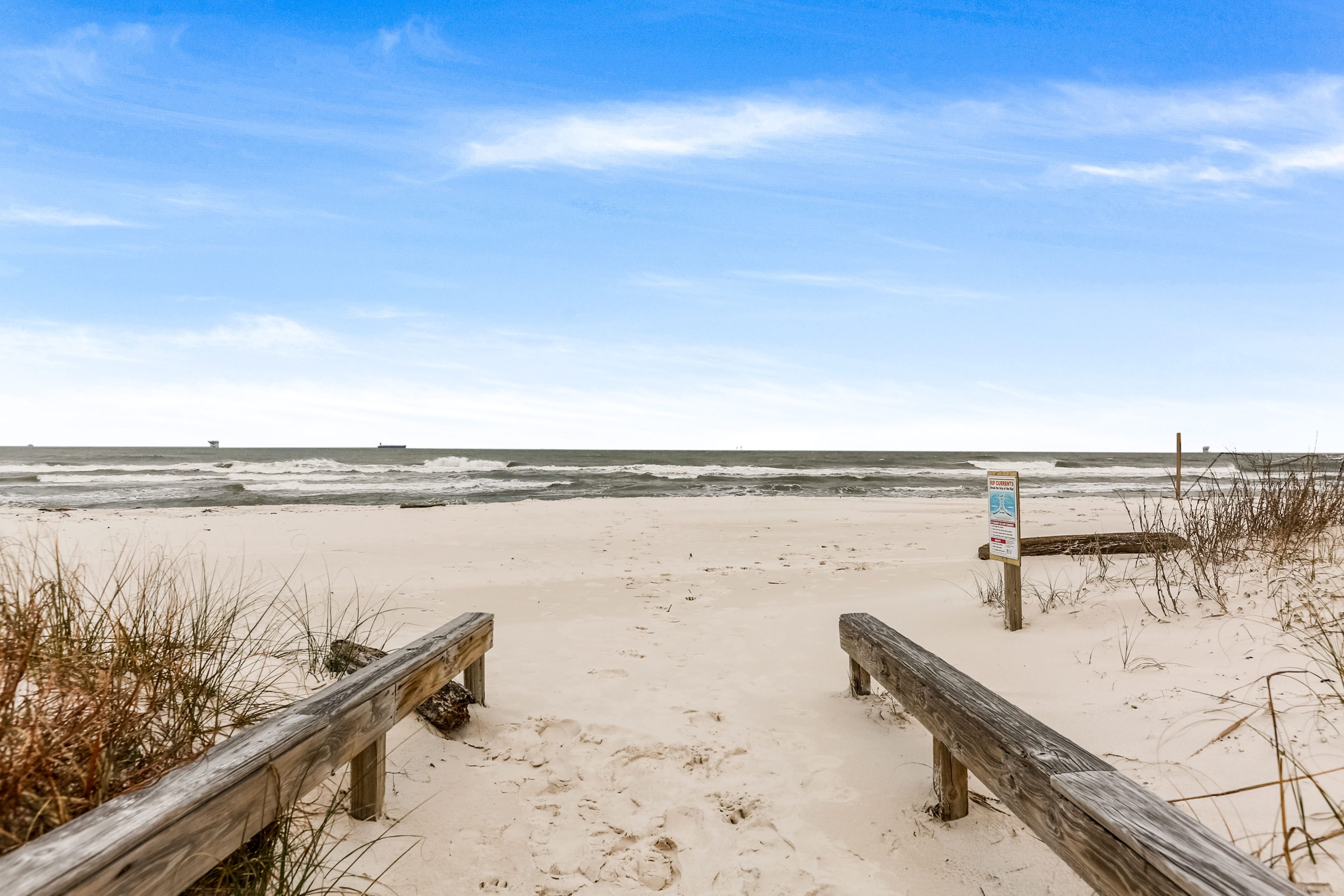Homokos strand, melynek mentén fa korlát és egy táblás útbaigazító található.