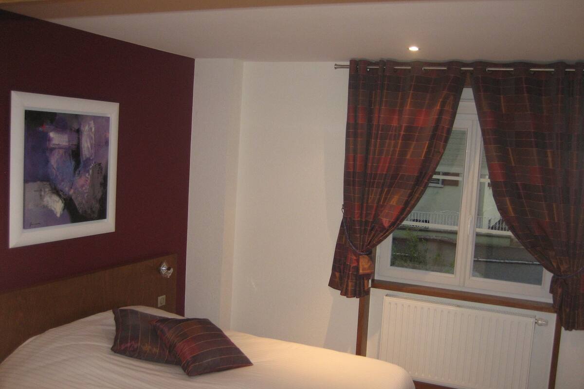 Ein Schlafzimmer mit Bett, einem Fenster mit Vorhängen, einem Heizkörper und einem gerahmten Bild an der Wand.