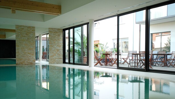 Una amplia piscina interior con paredes de vidrio transparente, vigas de madera en el techo y mobiliario exterior visible a través del vidrio.