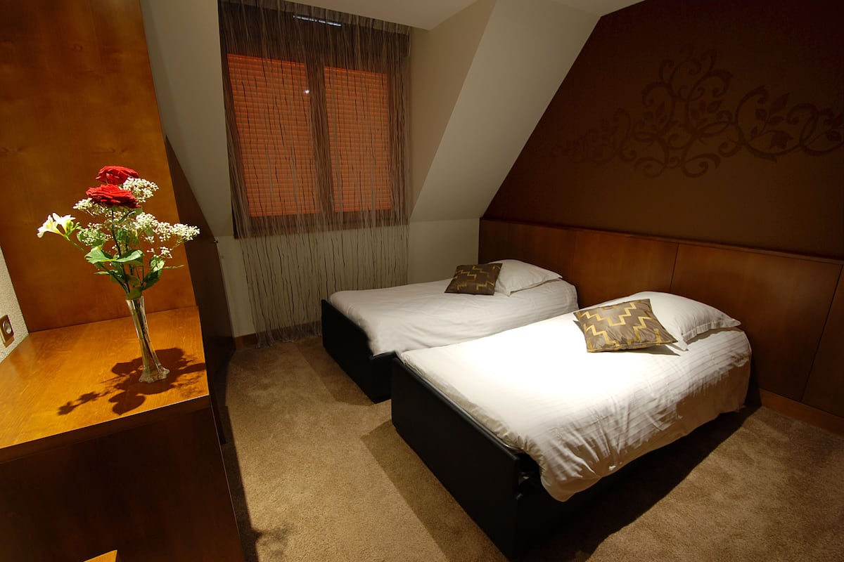 Ein Schlafzimmer mit zwei Betten, einem hölzernen Nachttisch, einer Vase mit Blumen und einem Fenster mit Jalousien.