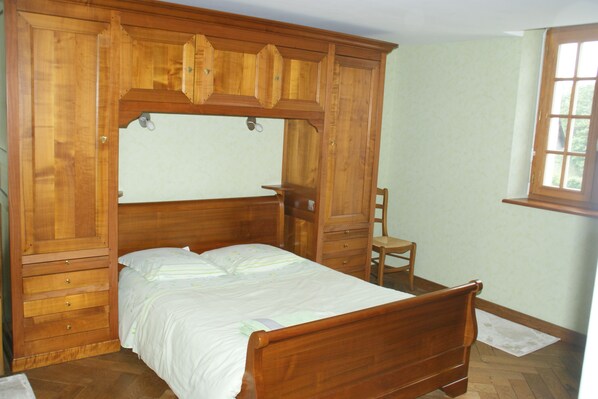 Una cama con dosel de madera, un cabecero, una pequeña silla de madera y una ventana con vistas a árboles.