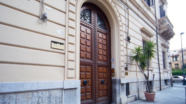 Una amplia puerta doble de madera con intrincados grabados, una planta en maceta a un lado y una fachada de edificio con base de piedra.