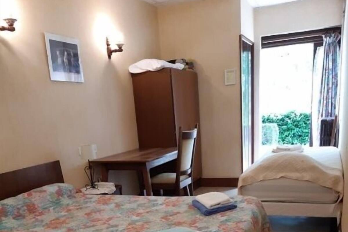 Une chambre d’hôtel avec un grand lit, un bureau et une chaise. Il y a une fenêtre avec des rideaux et une vue sur la verdure à l’extérieur.