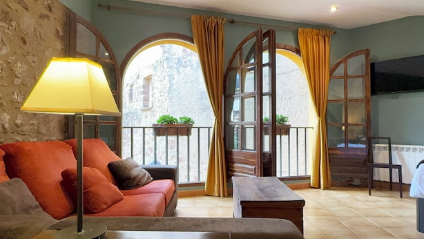 A cozy room with a red sofa, a small table, a floor lamp, and a TV on the wall. There are two windows with curtains and a balcony with plants. - Cal Crispí (Prades)