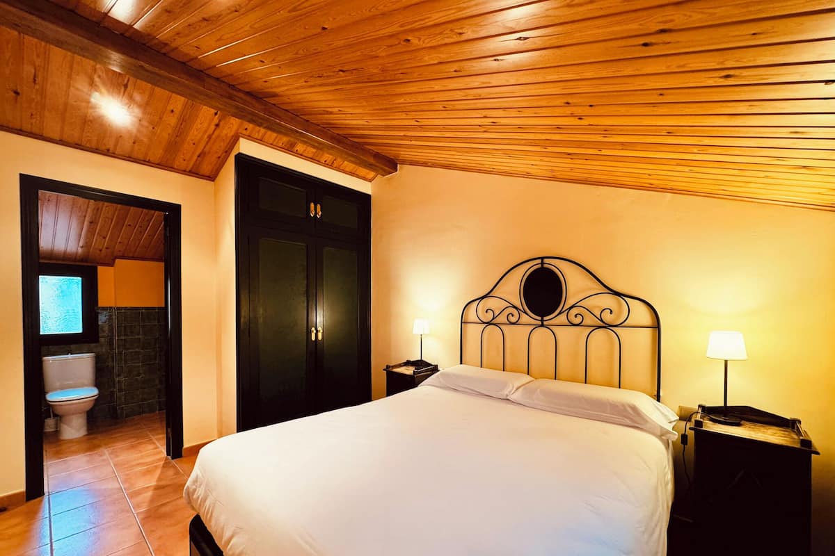 Une chambre d’hôtel avec un plafond en bois, un lit recouvert de draps blancs, une armoire noire et une salle de bain visible à travers une porte ouverte.