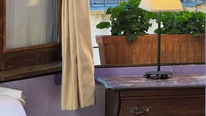 A room with a bed, a window with curtains, a lamp, and a view of a balcony with a railing and plants. - Cal Crispí (Prades)