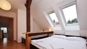 Quarto com teto inclinado, vigas de madeira, claraboia, cama com roupa branca e uma pequena mesa com um vaso.