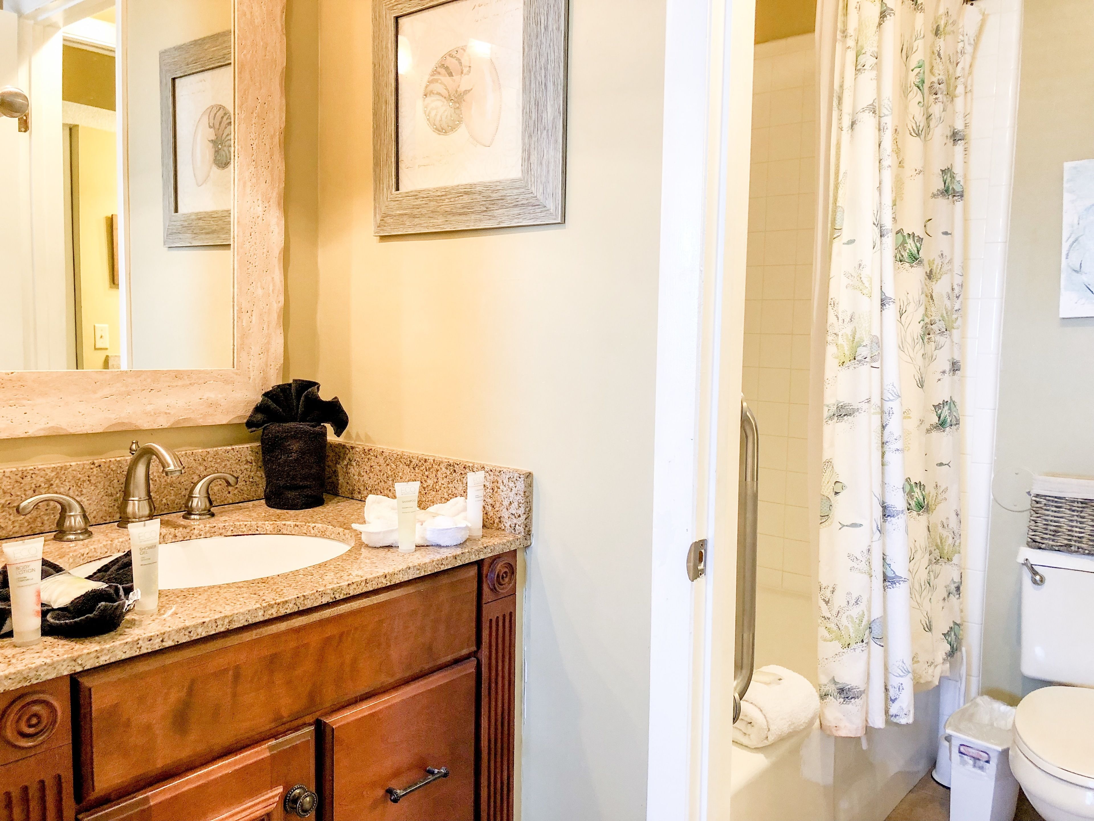 A bathroom with a wooden vanity, two sinks, a mirror, framed artwork, and a shower with floral curtains.