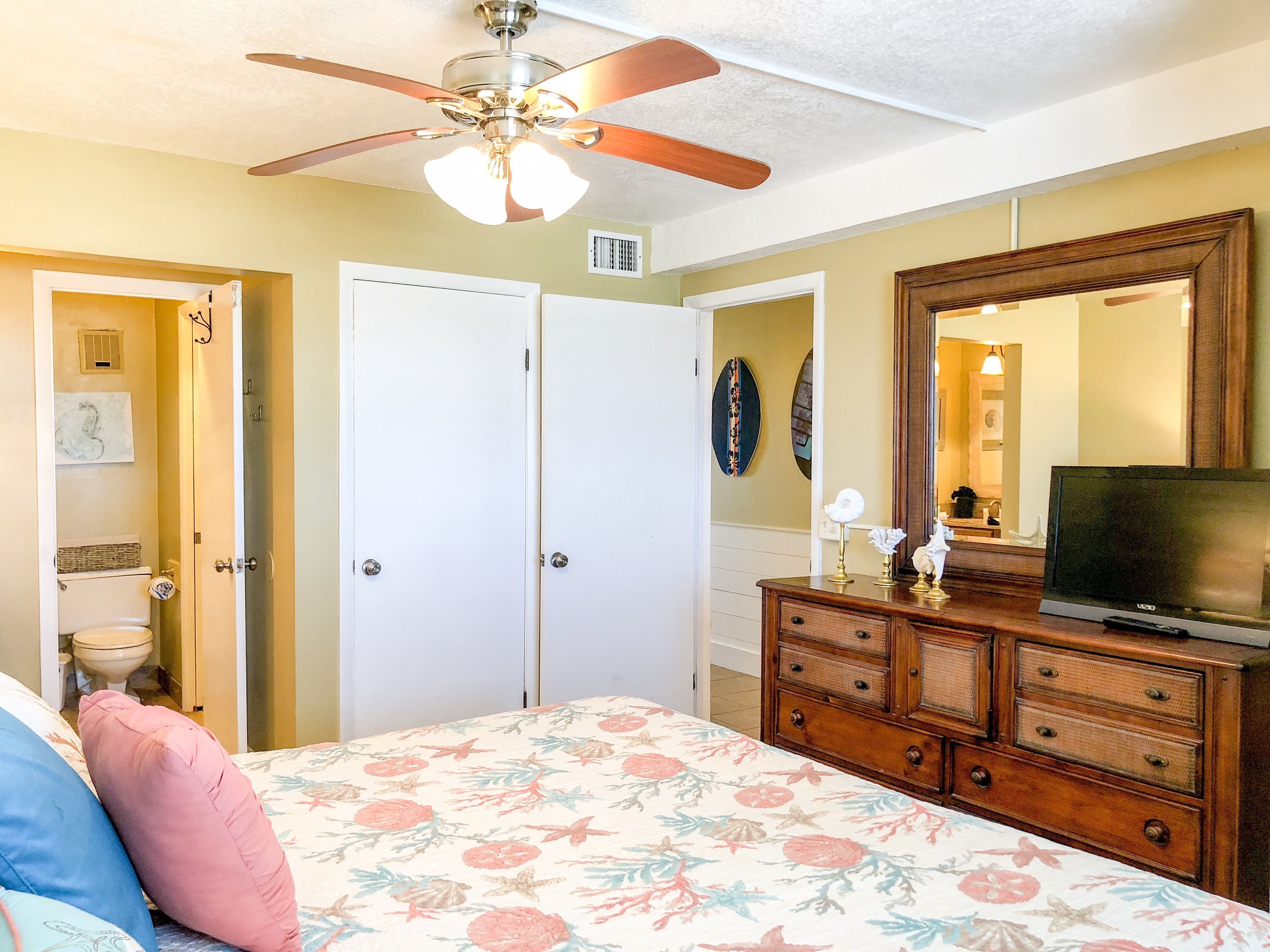 A bedroom with a bed, dresser, TV, ceiling fan, and a door leading to a bathroom.