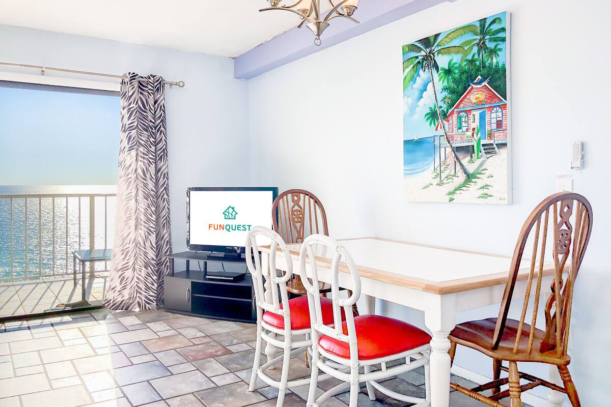 Een eethoek met een tafel en stoelen, een televisie waarop 'FUNQUEST' wordt getoond, een schilderij van een strand en een balkon met uitzicht.