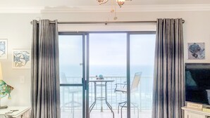 A room with a sliding glass door leading to a balcony with a view of the ocean. There is a television on the right side of the room. - Summit Beach Resort by H2O VR (Panama City Beach)