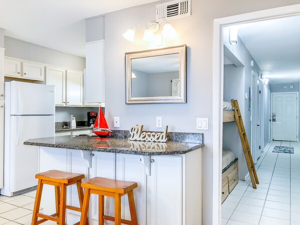 A kitchen with white cabinets, a granite countertop, two wooden stools, a red sailboat decoration, and a 'Welcome' sign.