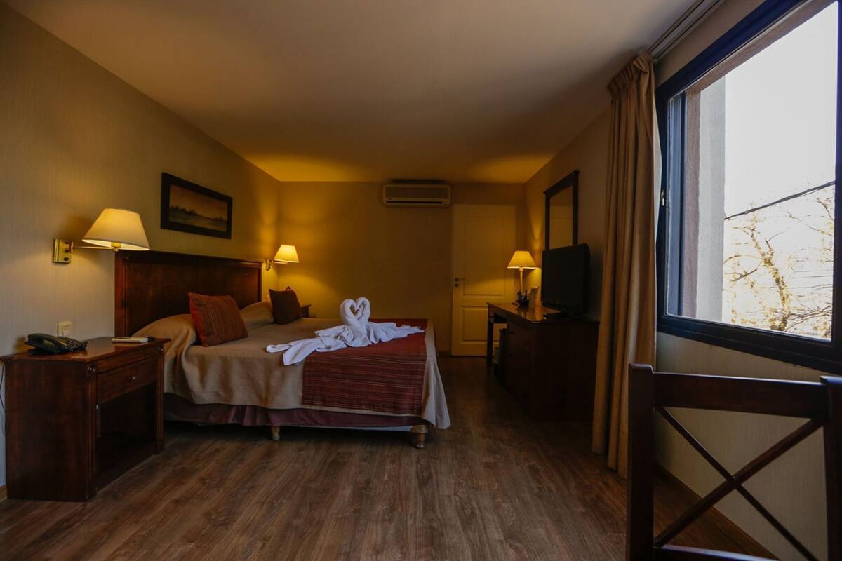 A hotel room with a bed, bedside lamps, a TV, a desk, and a window.