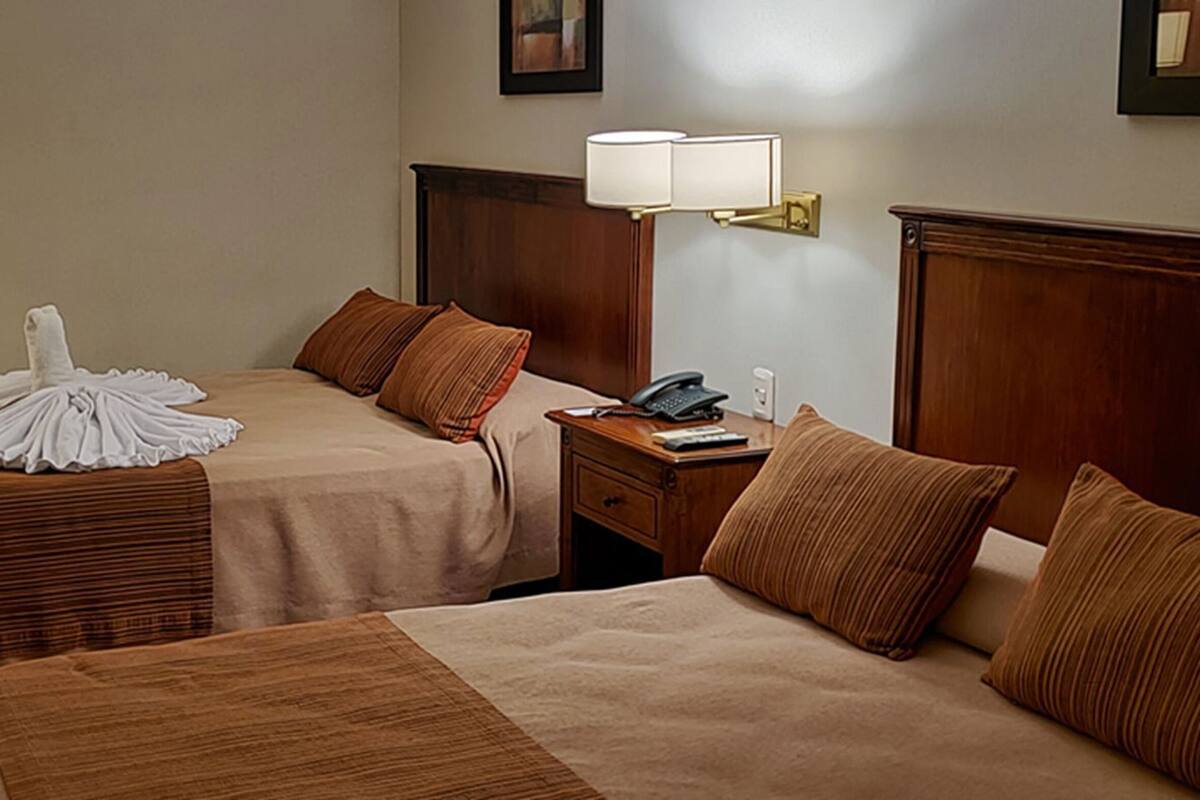 A hotel room with two beds, a nightstand, a lamp, and a chair.