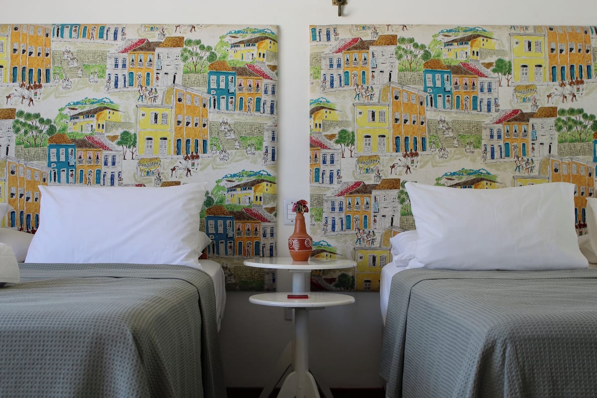 A hotel room with two beds, a small table with a vase, and colorful wall art depicting buildings and people.