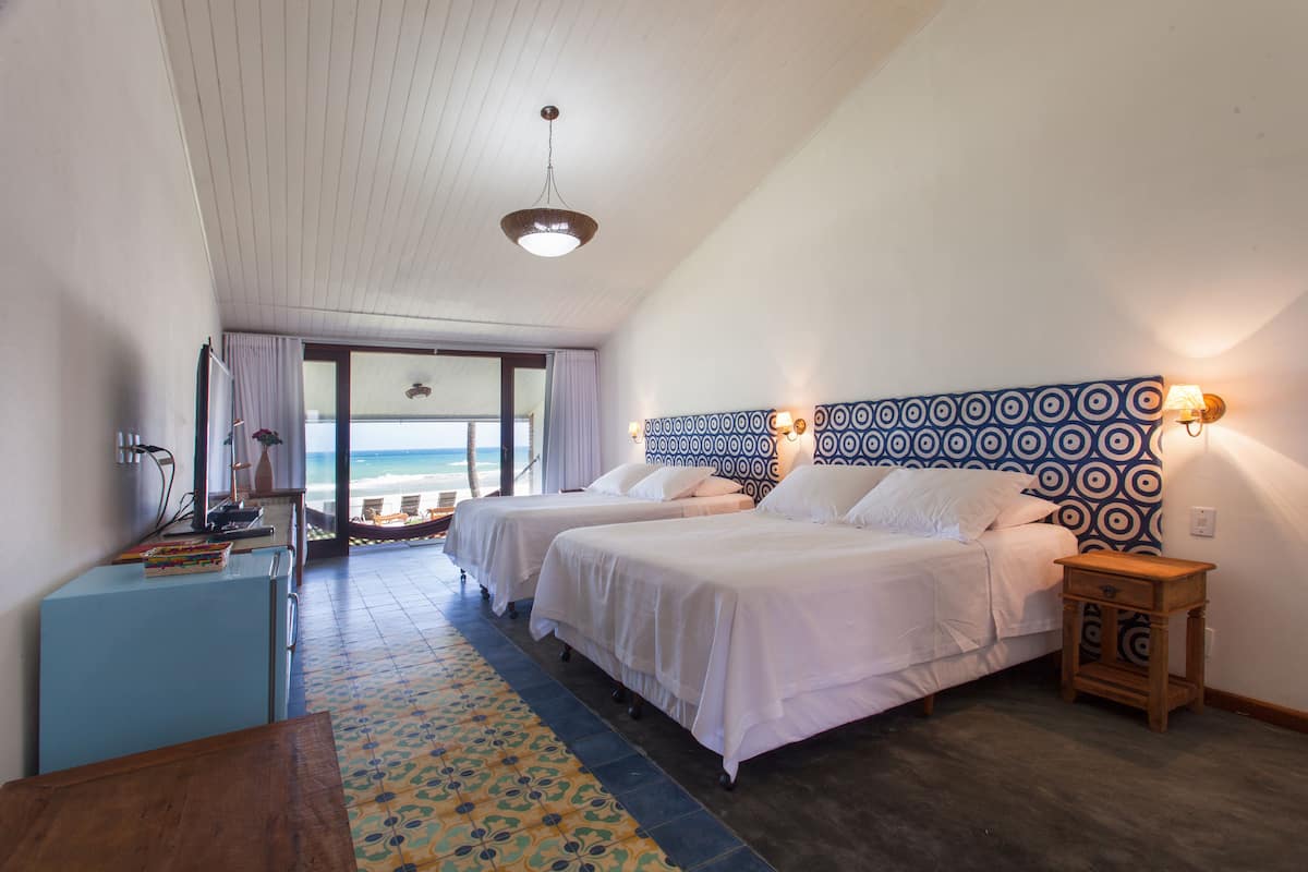 A hotel room with two beds, a wooden desk, a patterned rug, and a view of the beach.