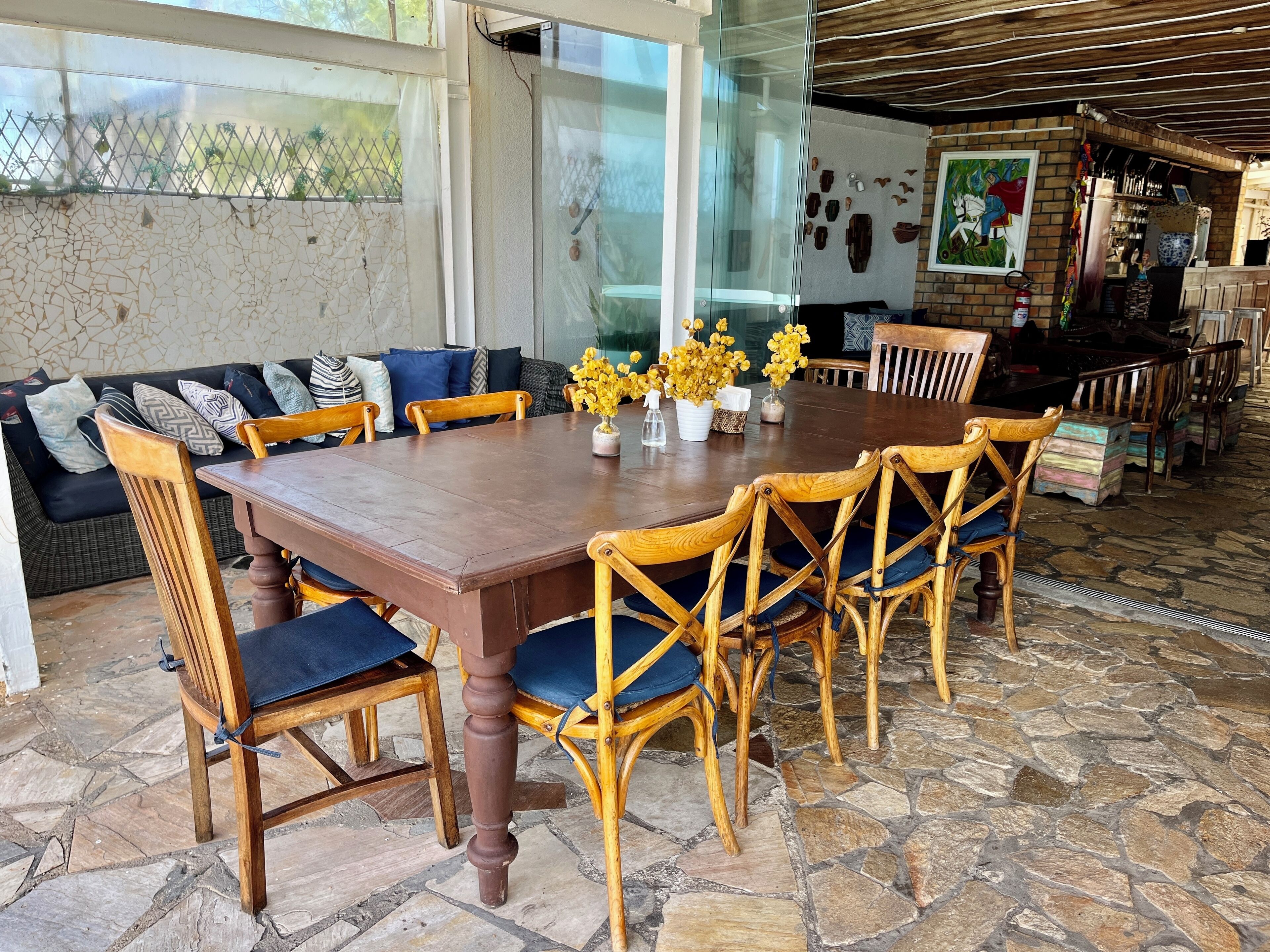 Un comedor con una gran mesa de madera rodeada de sillas de madera con cojines azules, una partición de vidrio y un piso de baldosas de piedra.