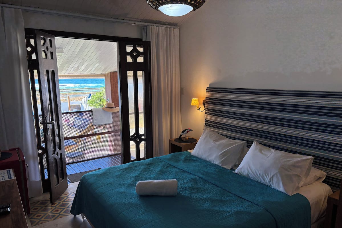 A hotel room with a bed, a nightstand, a lamp, and a view of the ocean through a door.