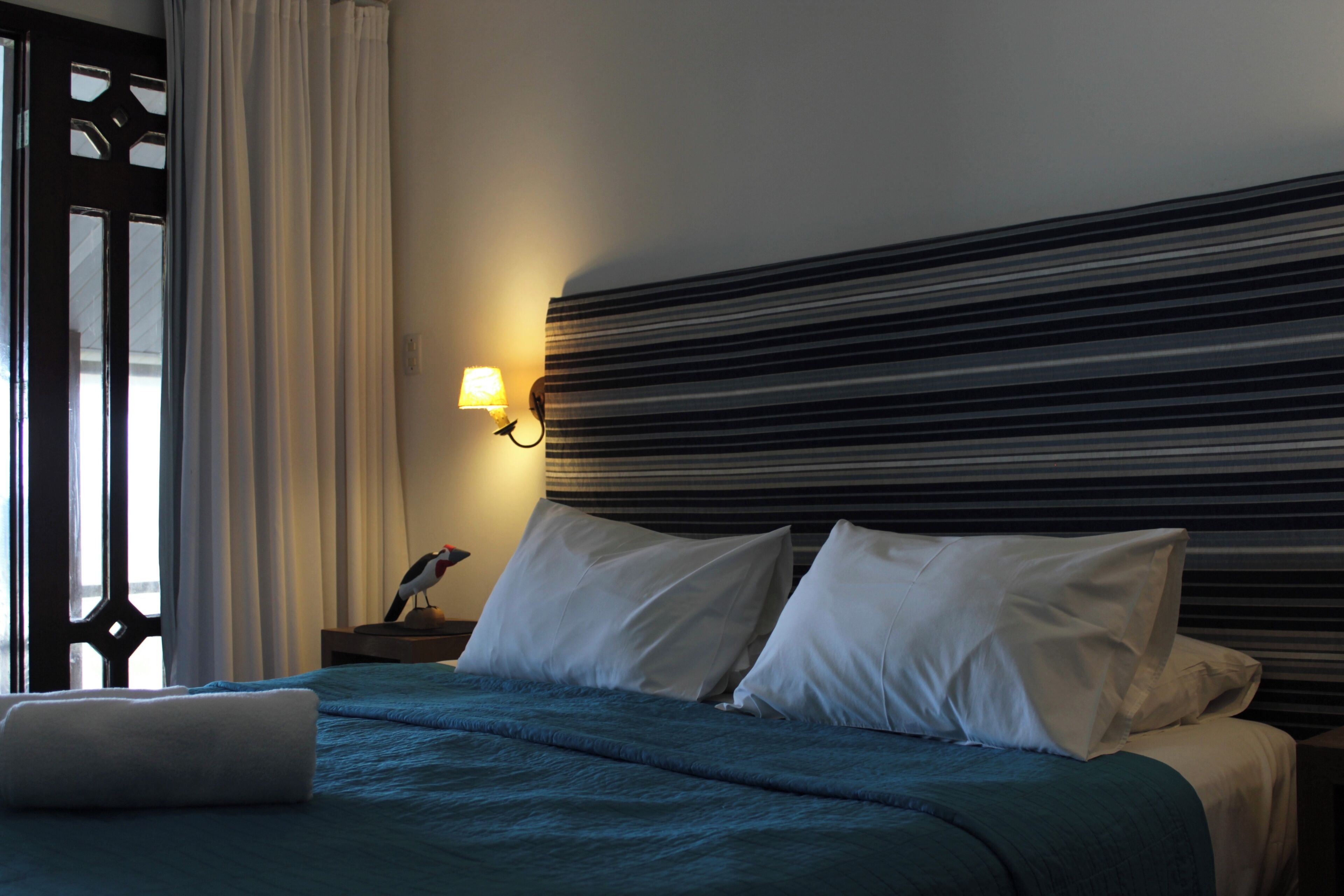 Habitación de hotel con cama, almohadas, mesita de noche, lámpara y ventana con cortinas.