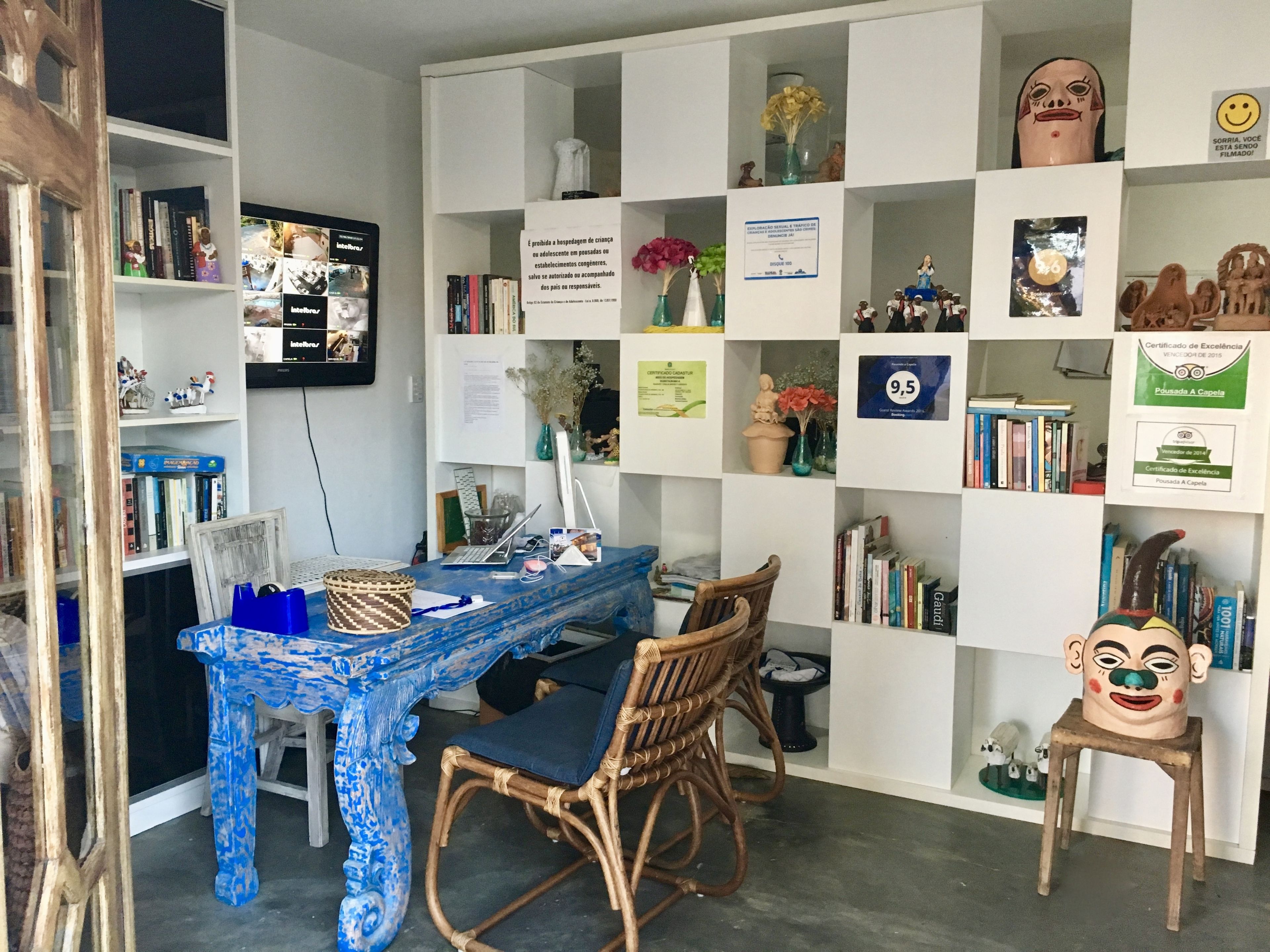 Une salle d’étude avec une table peinte en bleu, des chaises en osier, une bibliothèque remplie de livres et de certificats, et un téléviseur accroché au mur.