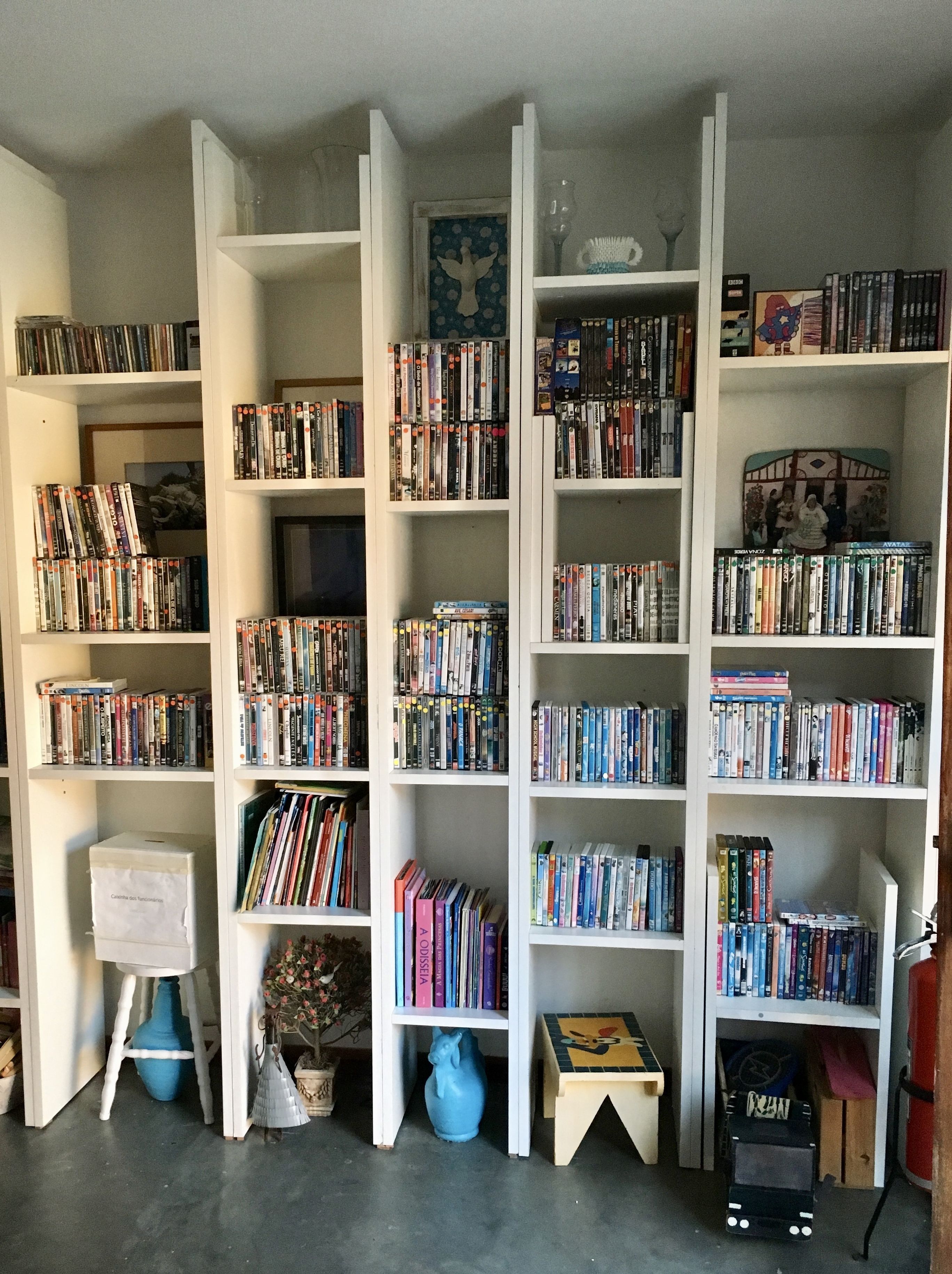 A room with white shelving units filled with books and DVDs. There is a small table with a vase and a chair.