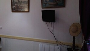 Una habitación con dos cuadros enmarcados en la pared, un televisor negro montado, un ventilador de pie y un radiador.
