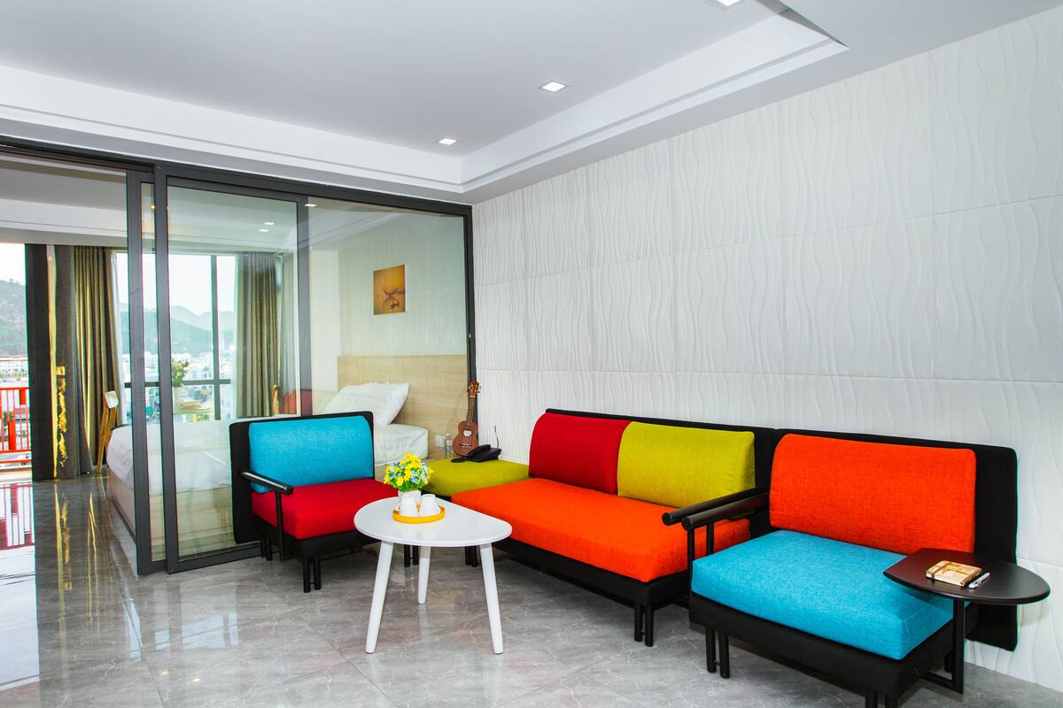 A modern hotel room with a colorful sectional sofa, a small white table with a vase, and a glass door leading to a balcony.