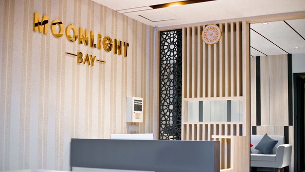 A hotel reception area with a modern design, including a reception desk, a decorative wall with the hotel's name, and a seating area with a sofa and a chair. - Moonlight Bay Hotel (Bac Nha Trang)