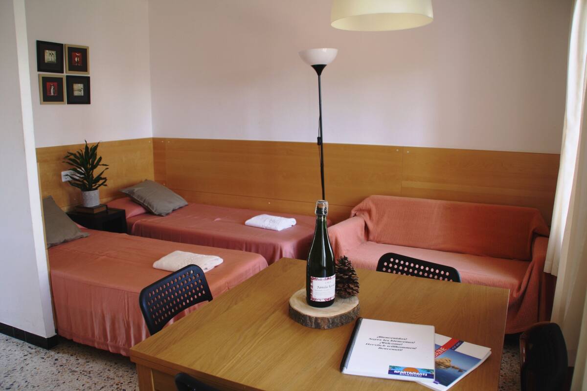 Habitación de hotel con dos camas individuales, una mesa de madera con una botella y una piña, una silla y una lámpara.
