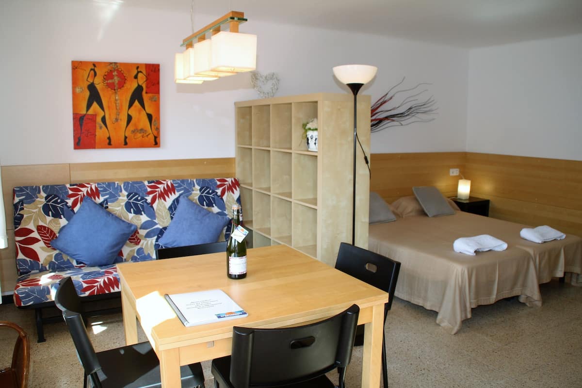 Una habitación de hotel con una mesa de madera, un sofá con cojines de colores, una cama con cubrecama color beige y una estantería.