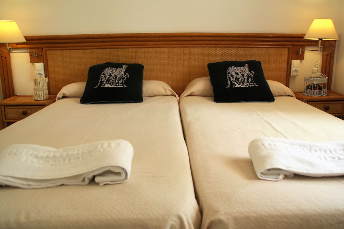 Dos camas individuales con ropa de cama color beige, almohadas negras con diseño blanco y toallas blancas dobladas al pie de cada cama.