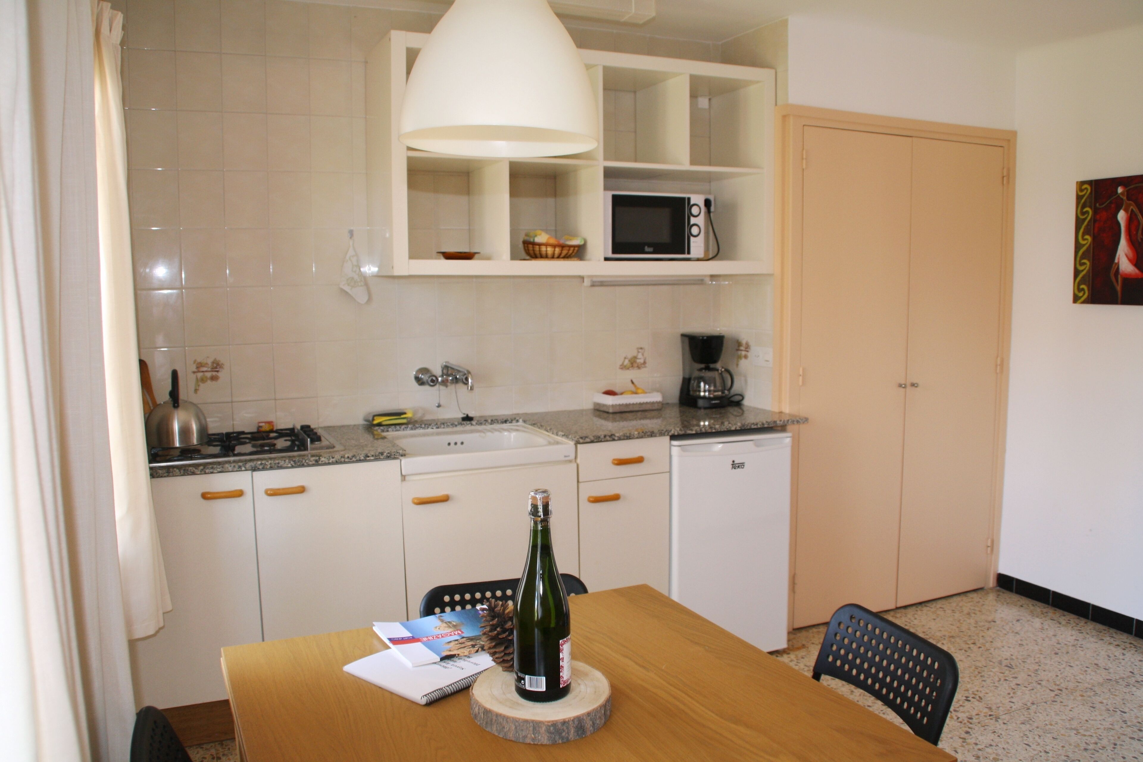 Een compacte keuken met witte kastjes, een magnetron, een koffiezetapparaat en een eettafel met een fles champagne.