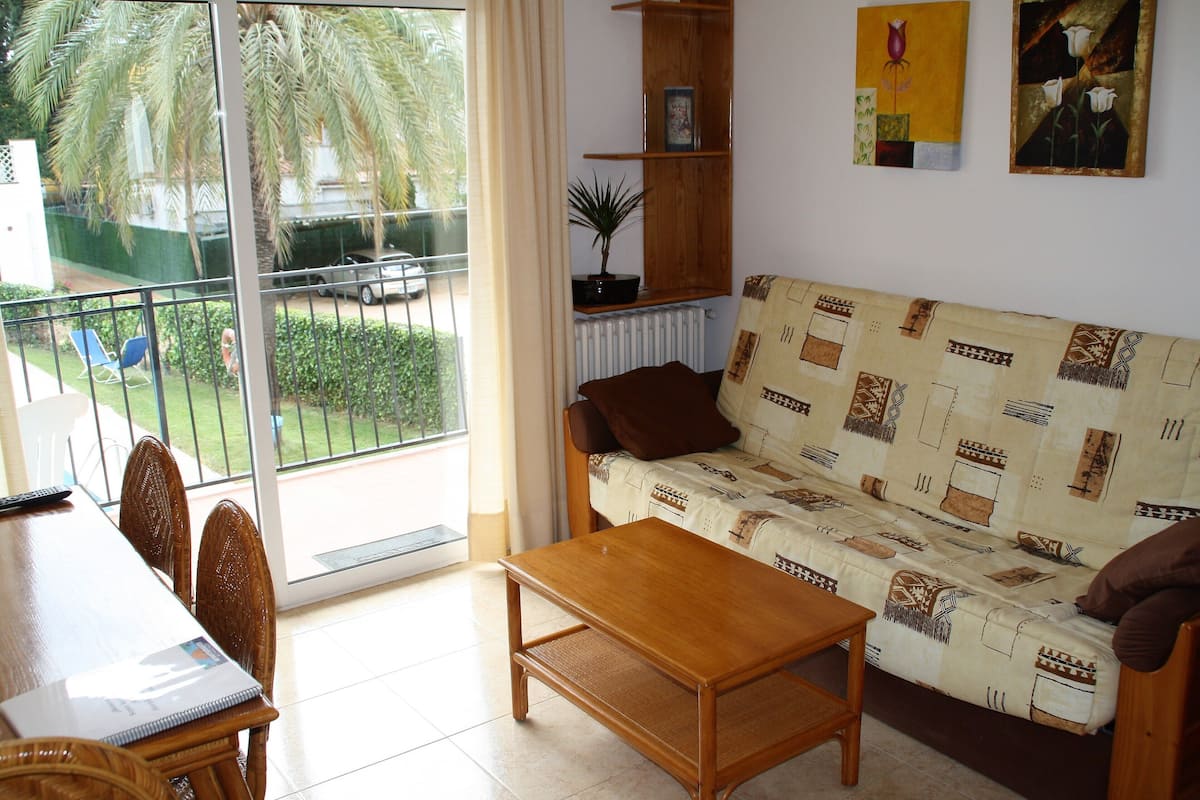 Una sala de estar con un sofá, una mesa de centro de madera y una mesa de comedor con sillas. Hay un balcón con vistas a palmeras y un coche.
