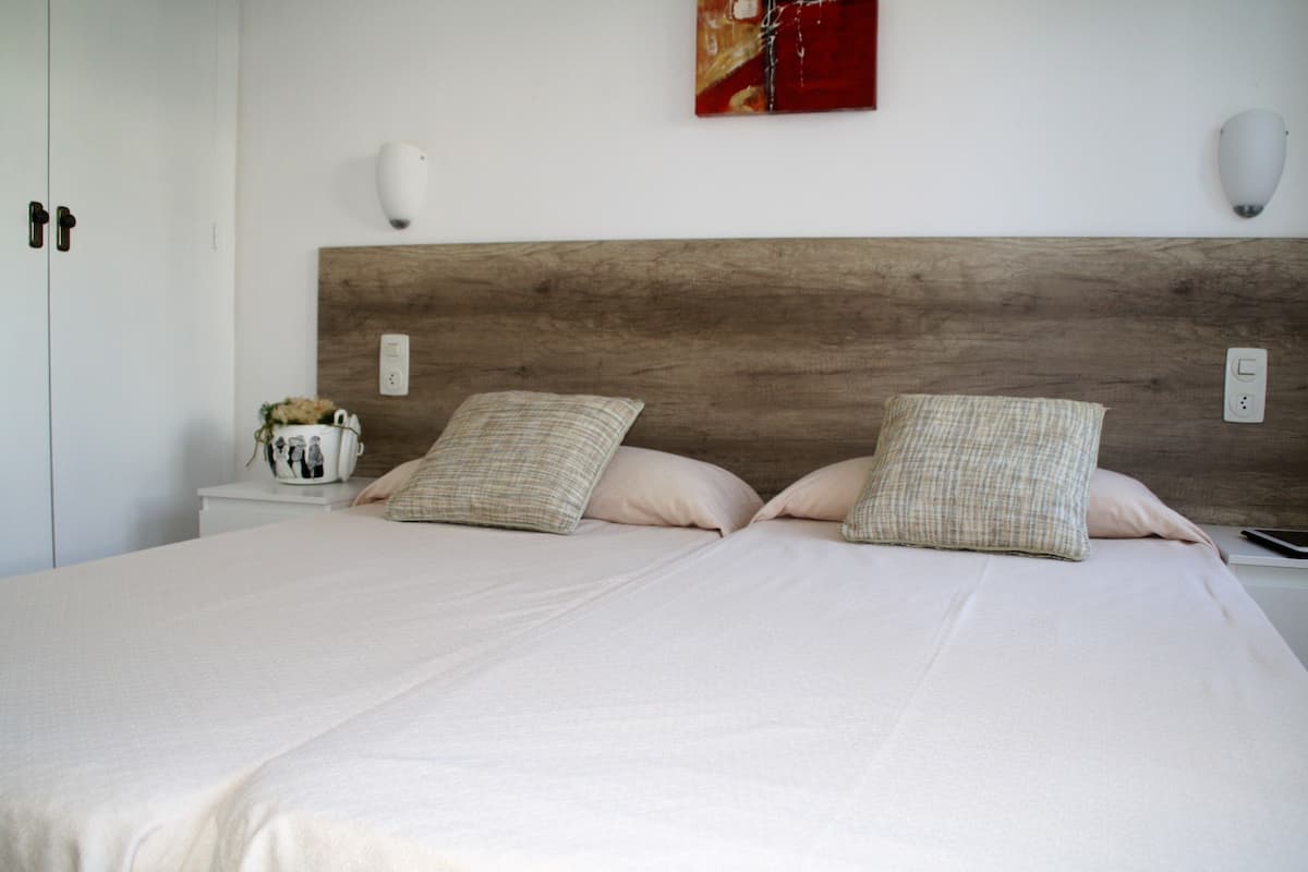 Un dormitorio con cabecera de madera, dos almohadas, una mesita de noche con un florero y una lámpara de pared.
