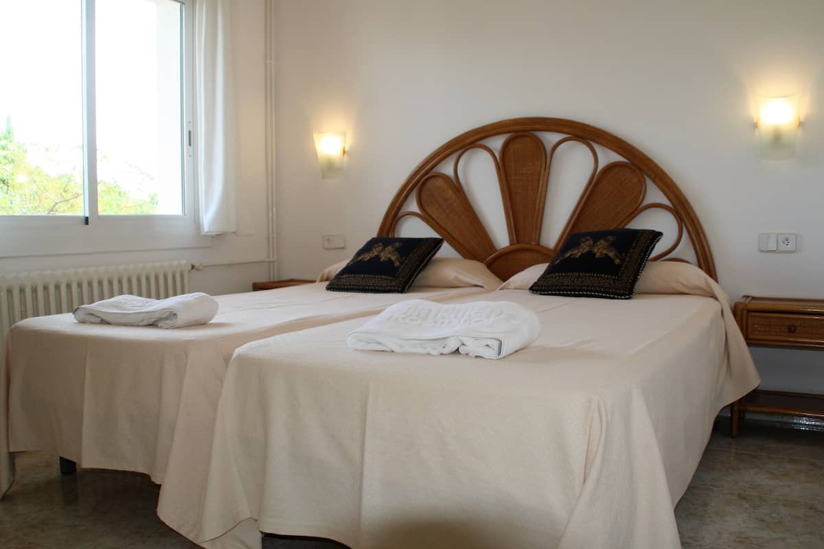 Habitación de hotel con dos camas, cada una con una colcha blanca y una toalla doblada. Hay lámparas de pared a ambos lados de las camas.