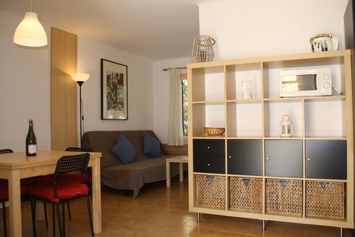 Una sala de estar con una mesa de comedor de madera, sillas rojas, un sofá con cojines azules, una estantería y un microondas en un estante.