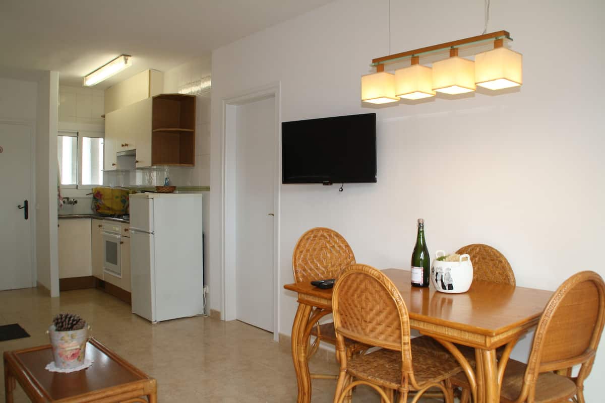 Un área de comedor con una mesa y sillas de madera, un televisor montado en la pared y una cocina al fondo.