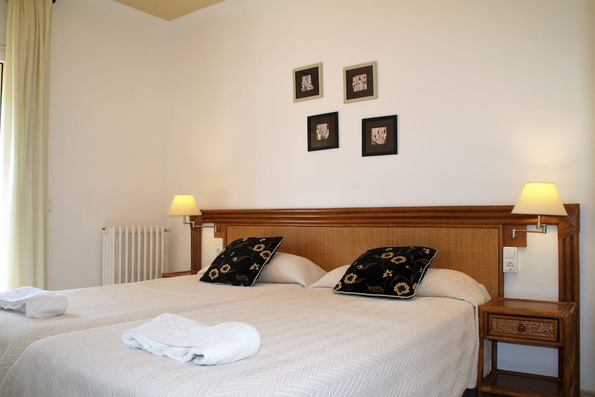Una cama bien hecha con una colcha blanca y dos almohadones decorativos, una mesita de noche con una lámpara y cuatro cuadros enmarcados en la pared.